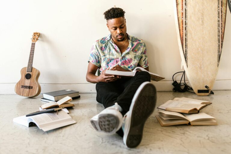 A young man reads a book while sitting among books and a ukulele, with a surfboard leaning on the wall.